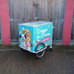 Turquoise dog ice cream cart with cartoon dogs and the text “Doggie Ice Cream for Dogs” on the side, parked on wet pavement in front of wooden barn doors.