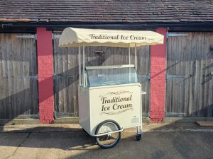 “A cream-colored traditional ice cream cart with a canopy labeled ‘Traditional Ice Cream’ and a glass display freezer. The cart has one large wheel and a small caster wheel and is positioned outdoors in front of wooden garage doors with red brick pillars in sunlight.” 🍦